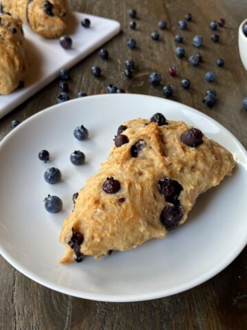 A Blueberry Scone on a small white plate in the foreground, additional scones and a bowl of blueberries in the background.