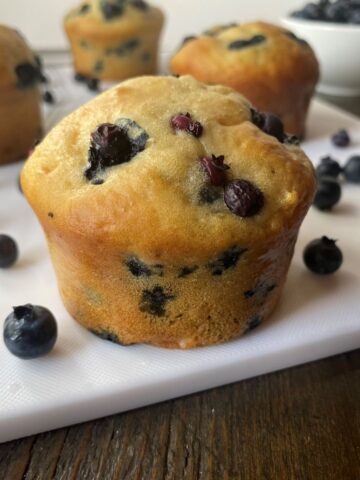 A Healthy Blueberry Muffin with blueberries scattered around it in the foreground and three blueberry muffins and a bowl of blueberries in the background.