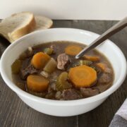 Slow Cooker Beef and Barley Soup in a white bowl in the foreground. Slices of bread in the background.