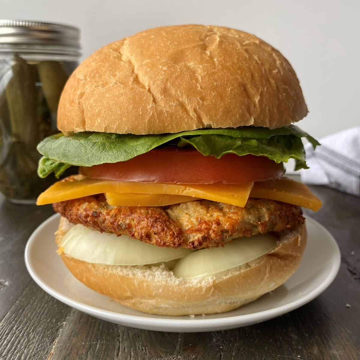 Chicken Burger with all of the fixings on a plate in the foreground, with a jar of pickles and dishtowel in the background.