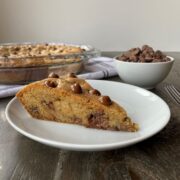 A piece of Chocolate Chip Cookie Pie on a white plate in the foreground, with the remaining cookie pie and a dish of chocolate chips in the background.