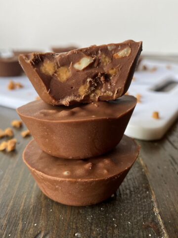 A stack of three Toffee Chocolate Almond Cups and toffee bits in the foreground and more Toffee Chocolate Almond Bites in the background.