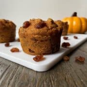A vegan pumpkin muffin with raisins sprinkled around it on a white cutting board in the foreground with two vegan muffins and a pumpkin in the background.