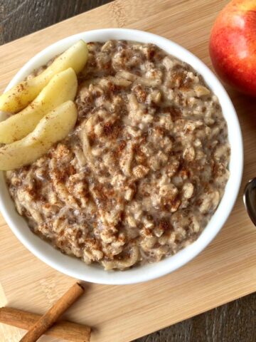 A bowl of oatmeal with sliced apples on top in the middle of the photo, with an apple, cinnamon sticks and a spoon surrounding the bowl.