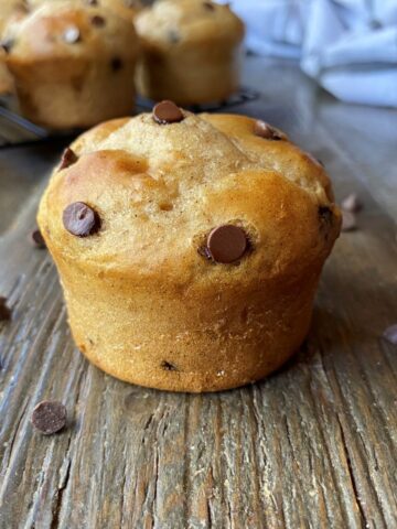 A chocolate chip muffin with chocolate chips scattered around the muffin in the foreground with chocolate chips muffins and a tea towel in the background.
