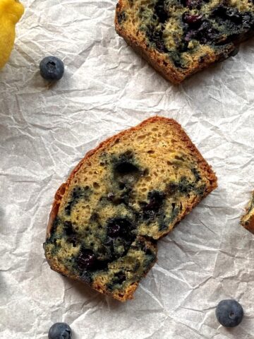A piece of lemon blueberry bread in the middle with part of the loaf on the right side and another piece of bread and a lemon on the top.