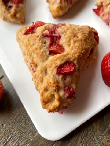 A strawberry scone on a white board with fresh strawberries beside the scone and other scones in the background.