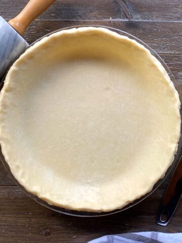 Pie crust in a glass pie plate with a rolling pin on the top left and a pie lifter on the right.