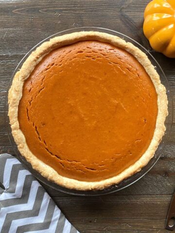 A baked pumpkin pie in the middle with an oven mitt on the left side and a pie lifter and pumpkin on the right side.