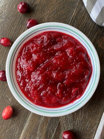 A bowl of cranberry sauce in the center surrounded by fresh cranberries and a metal spoon on the right side.