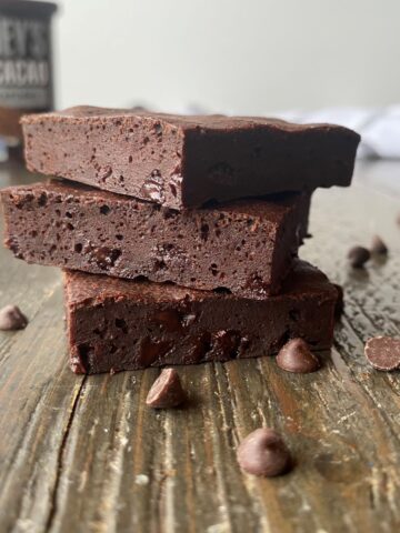 Three pieces of healthy brownies stacked on top of one another with mini chocolate chips surrounding the brownies and a container of cocoa powder in the left background.