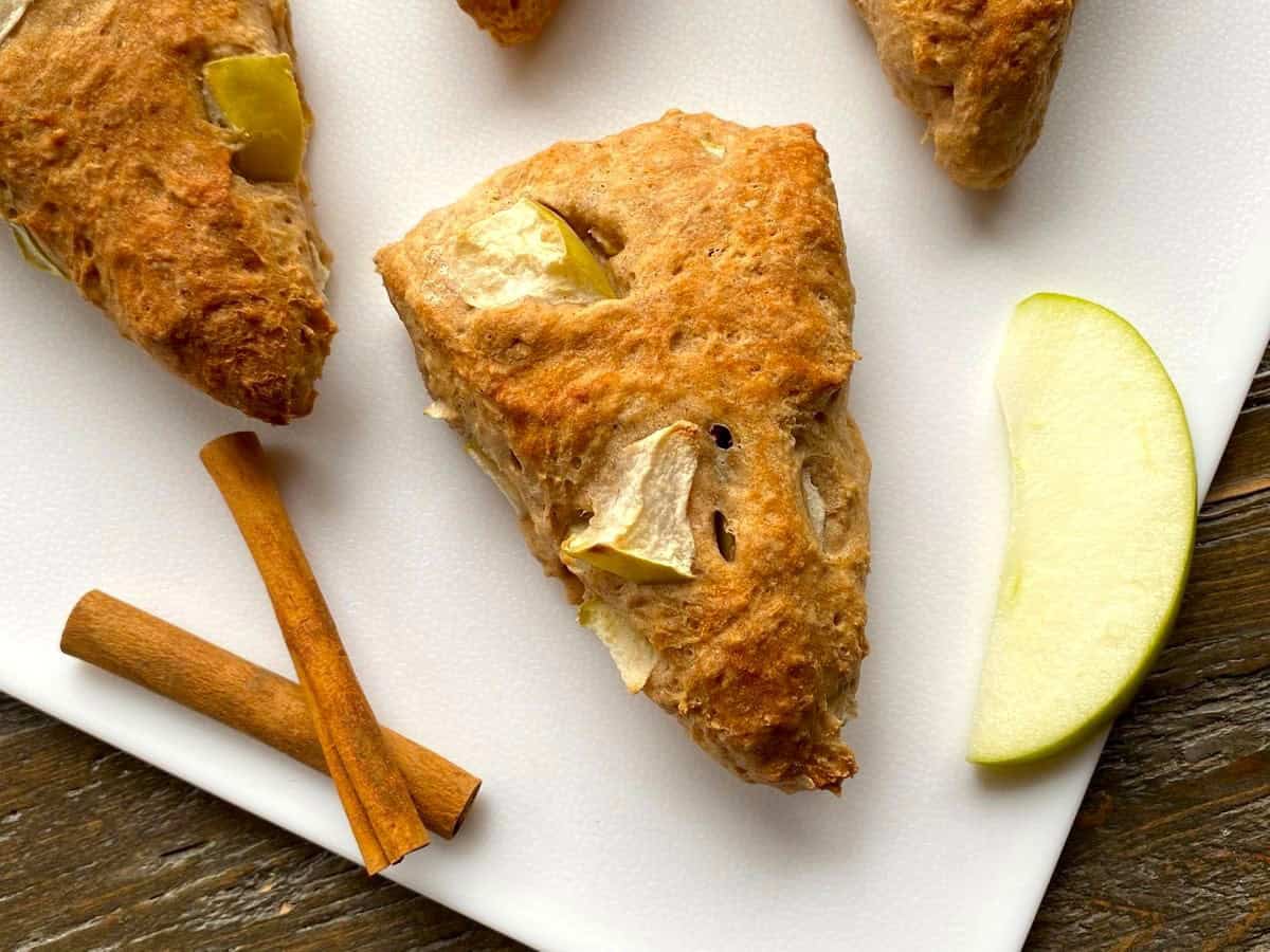 Baked scones with cinnamon sticks and an apple slice on a white cutting board.